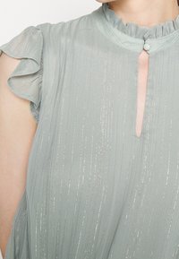 Ljusgrön blus med vertikala silverstrimmor, volang korta ärmar, hög krage med en knapp och en nyckelhålsöppning fram.