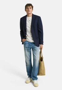 Blazer marine, camiseta blanca con logo, jeans azul desgastado, zapatillas crema y un bolso de ante beige. Conjunto informal con elementos texturizados.