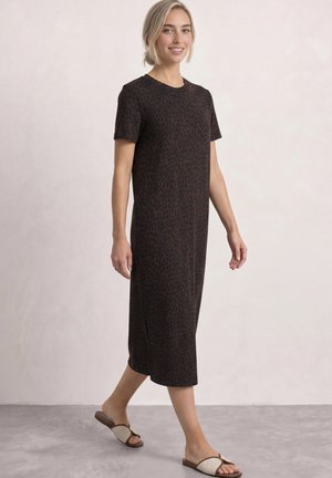 Femme aux cheveux blonds courts marchant, portant une robe midi marron foncé et noire à motifs et des sandales plates beiges à bout ouvert, souriant légèrement.