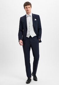 Marineblauer Anzug mit einem weißen Hemd und einer hellgrauen Weste, ergänzt durch eine Fliege. Schwarze Lederschuhe runden das formelle Outfit ab.