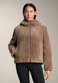 Brun fuskpälsjacka med hög krage, dragkedja fram och långa ärmar. Mjuk textur och avslappnad passform, ihop med svarta leggings.