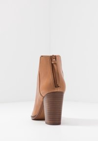 Stivale alla caviglia in pelle color cuoio con una forma curva, cerniera posteriore e tacco a blocco con scanalature orizzontali. Texture liscia nel complesso.