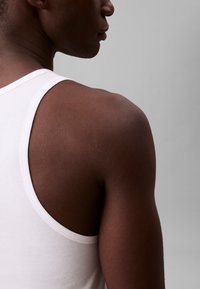 Joven con camiseta de tirantes blanca mostrado de hombro y perfil lateral contra fondo gris liso.