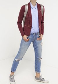 Blazer bordeaux sopra una camicia azzurra chiara, jeans blu strappati e sneakers grigie. Zaino argentato con spalline regolabili. Stile classico.