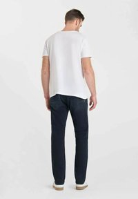 T-shirt blanc à manches courtes avec un col arrondi, associé à un jean bleu foncé et des baskets blanches. Le t-shirt a un dos légèrement allongé.