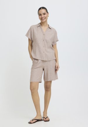 Mujer sonriendo, vestida con camisa beige de manga corta con botones y pantalones cortos a juego hasta la rodilla con rayas verticales blancas, de pie con las manos en los bolsillos.