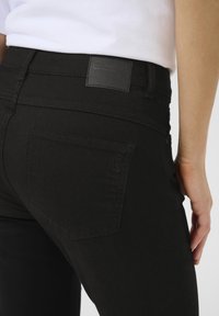 Čierne denimové nohavice so slim fit strihom, s jednou zadnou kapsou a štruktúrovanou koženou nášivkou na páse. Hladká tkanina.