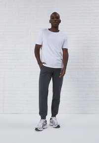 Weißes T-Shirt mit kurzen Ärmeln kombiniert mit dunkelgrauen Jogginghosen und silbernen Sportschuhen. Das Model nimmt eine entspannte Pose vor einem weißen Backstein-Hintergrund ein.