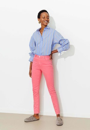 Femme souriante aux cheveux courts et bouclés, portant une chemise bleue à rayures boutonnée, un pantalon rose et des chaussures slip-on à imprimé léopard.