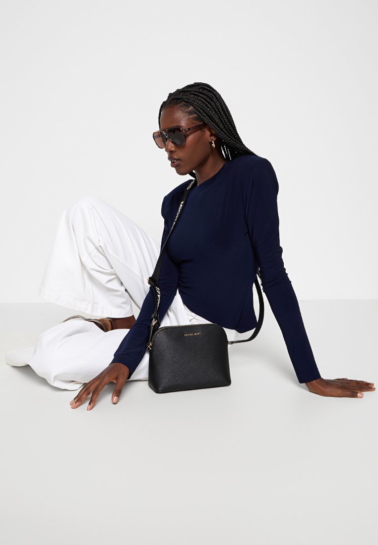 Schwarze Ledertasche mit strukturierter Form, goldenen Beschlägen und verstellbarem Tragegurt. Kombiniert mit einem marineblauen Langarmshirt und weißen Hosen.