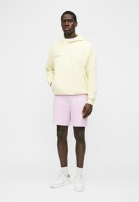 Mand iført en lysegul hættetrøje, lyserøde shorts, hvide sokker og hvide sneakers, der står foran en ensfarvet hvid baggrund.