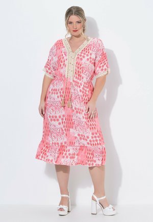 Frau trägt ein knie- bzw. knielanges Kleid mit pink-weißem Muster und Fransen, steht in weißen Sandalen mit hohen Absätzen vor weißem Hintergrund.