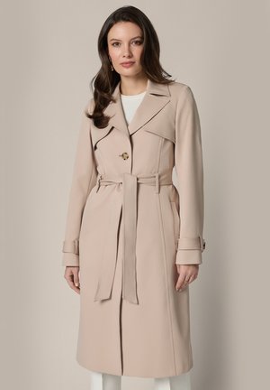 Kobieta o długich brązowych włosach ubrana w beżowy trencz z paskiem na białej bluzce, stojąca na neutralnym tle.