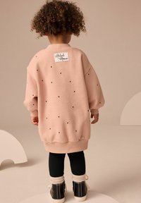 Barn med lockigt hår klädd i en överstor, ljusrosa klänning med svarta hjärtan och Minnie Mouse-märke, svarta leggings och svarta stövlar som står inomhus.