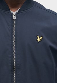 Tengerészkék bombázó dzseki sima anyagból, ezüst cipzárral és arany hímzett madár logóval a bal mellrészen.