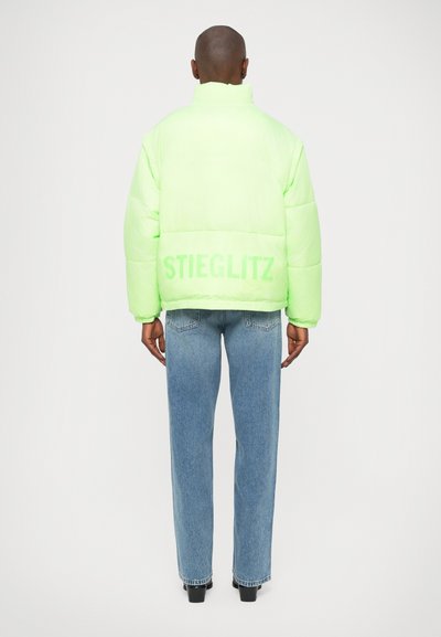 Neon zelena pufasta jakna s visokim ovratnikom, s logotipom "STIEGLITZ" na leđima, kombinirana s svijetloplavim ravnim trapericama.