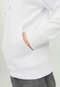 Haut à capuche blanc en coton avec une poche kangourou, des poignets et un ourlet côtelés. Texture lisse avec des cordons de serrage et des accents métalliques sur la capuche.
