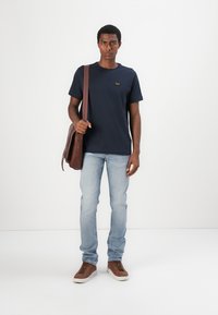 T-shirt en coton bleu marine avec un petit logo, jeans en denim bleu clair, sac messager en cuir marron, baskets marron, debout sur un fond blanc.