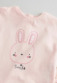 Camisa de algodón rosa claro con una cara de conejo y acentos de costura rosa. El diseño incluye el texto "Sonríe" y detalles de rayas en el interior de las orejas.