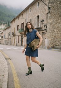 Femeie în rochie din denim care merge pe o stradă cu clădiri din piatră, ținând o geantă mare din piele întoarsă și purtând șosete verzi cu pantofi negri.