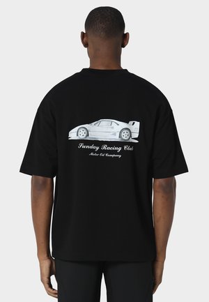Uomo che indossa una maglietta nera con una grafica bianca di un'auto sportiva e il testo "Sunday Racing Club Motor Oil Company" sulla schiena, visto di spalle.