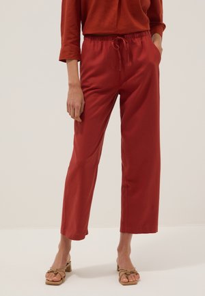 Persona che indossa pantaloni rossi larghi con coulisse, un top rosso coordinato con maniche e sandali color cuoio, in piedi contro uno sfondo chiaro e semplice.