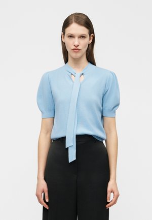 Femme portant un chemisier bleu clair à manches courtes avec un nœud sur le devant et un pantalon noir taille haute, debout devant un fond uni.