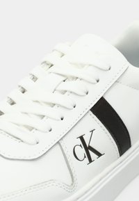 Λευκό δερμάτινο αθλητικό παπούτσι με μαύρη ρίγα, στρογγυλή μύτη, επίπεδα κορδόνια και διακριτικό λογότυπο. Ομαλή υφή με ραμμένες λεπτομέρειες.