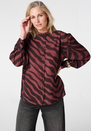 Blonde vrouw die een bordeauxrode en zwarte blouse met zebraprint, pofmouwen en donkerblauwe spijkerbroek draagt, poserend met haar hand vlak bij haar gezicht.
