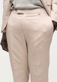 Pantalon beige sur mesure avec passants de ceinture, porté avec un haut léger en tricot et un blazer beige assorti sur une personne à la peau foncée, la main gauche visible.