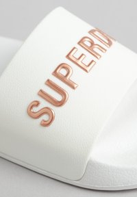 Sandalia deslizante blanca con una superficie texturizada, que presenta un prominente logo en oro rosa "SUPERDRY" en la correa. Diseño simple y minimalista.