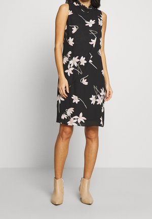 Ærmeløs sort knælang kjole med lyserødt blomsterprint båret med beige ankelstøvler på neutral baggrund.