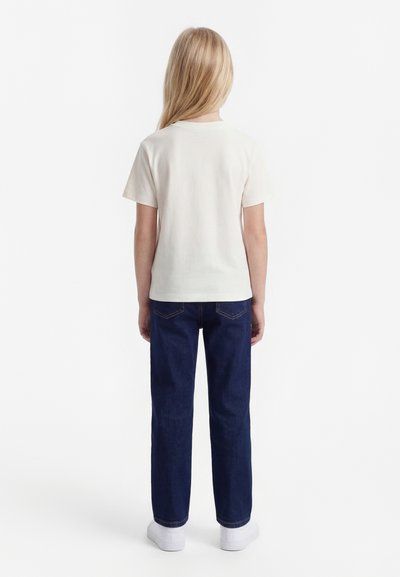 Copil cu păr blond lung, văzut de spate, purtând un tricou alb simplu cu mâneci scurte, blugi albastru închis și adidași albi.