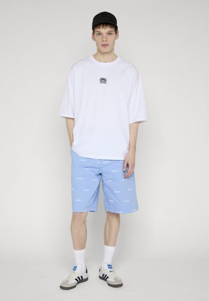 Noor mees, kes kannab valget ülikõrget T-särki, helesiniseid mustrilisi lühkareid, valgeid sokke, tenniseid ja musta mütsi, seisab ühevärvilise tausta ees.