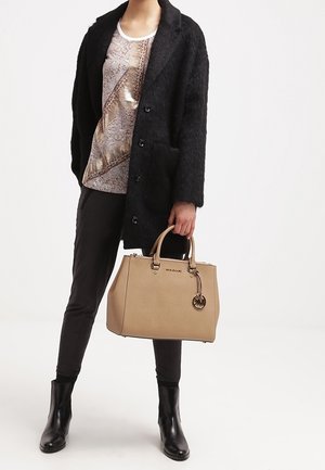 Personne portant un manteau noir, un haut beige à motifs, un pantalon noir, des bottes noires, tenant un sac à main beige Michael Kors avec un pendentif en forme de logo.