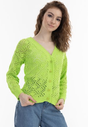 Vrouw met krullend haar die een felgroene gebreide knoopvest en blauwe spijkerbroek draagt, poserend met handen bij de zakken tegen een witte achtergrond.