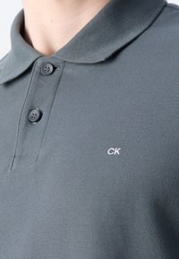 Polo gris en tissu texturé, avec un col classique, deux boutons et un petit logo blanc sur la poitrine.