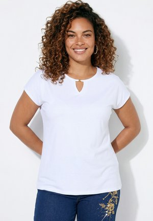 Frau mit lockigem Haar, die lächelt, trägt ein weißes Kurzarmshirt mit Schlüssellochausschnitt und blaue Jeans mit goldener Blumenstickerei.