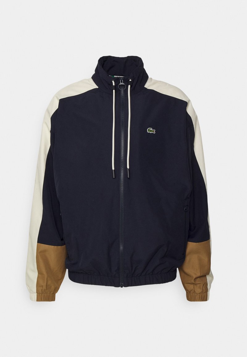 Marine windbreaker jas met beige en bruine accenten, volledige rits, staande kraag, elastische manchetten en zoom, met een klein logo op de borst.