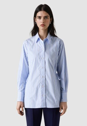 Młoda kobieta o ciemnych włosach ubrana w jasnoniebieską i białą koszulę w pionowe paski oraz ciemne spodnie, stoi na tle jednolitego tła.