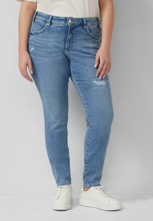 Osoba ubrana w jasnoniebieskie obcisłe jeansy z delikatnym przetarciem oraz białe sneakersy, stojąca na jednolitym szarym tle.