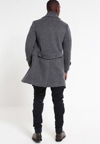 Abrigo de espiga gris con un corte ajustado, diseño de un solo pecho y detalles abotonados; combinado con pantalones negros de corte slim y botas hasta el tobillo.