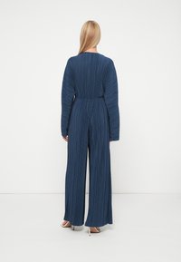 Tumši zila plisēta kombinezona ar apaļu dekoltē un garām piedurknēm, ar ietītu jostasvietu un platām biksēm. Gluda, plūstoša auduma.