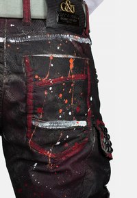 Zwarte denim jeans met spetterverf in oranje, wit en rode tinten. Zijzak met textuuraccenten en leren merkembleem op de taille.