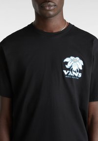 Camiseta de algodón negra con un gráfico de flor blanca con acentos azules y texto "VANS" en la parte izquierda del pecho. Mangas cortas y cuello redondo.