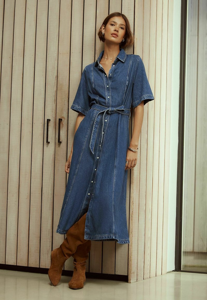 Femeie sprijinită de un perete de lemn, purtând o rochie-cămășă din denim cu curea și cizme maro din piele întoarsă până la genunchi, cu păr scurt și bijuterii minimaliste.