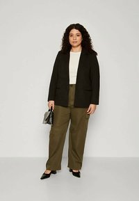 Sieviete ar burzglainiem matiem, ģērbta melnā žaketē, baltā topā, olīvu krāsas cargo biksēs, melnās augstpapēžu kurpēs, tur mazu melnu rokassomu.