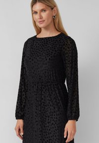 Kobieta z długimi blond włosami, ubrana w czarną, przezroczystą suknię z długimi rękawami, w fakturę groszków, z srebrnymi kolczykami w kształcie kół.