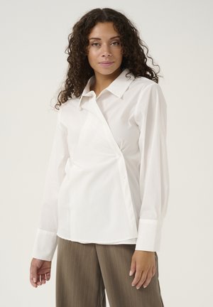 Camicia bianca con maniche lunghe, caratterizzata da un design asimmetrico unico e una texture liscia. Abbinata a pantaloni marroni a righe. }}
