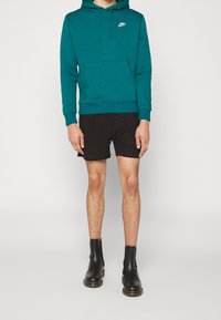 Tyrkysová bavlněná mikina s přední kapsou a stahovací kapucí, spárovaná s černými šortkami a černými kotníkovými botami s hladkým povrchem.